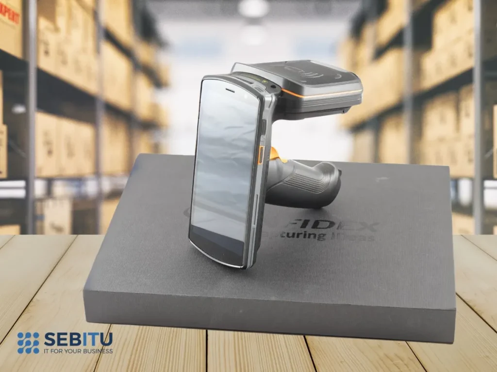 Mobilny terminal RFID z ekranem dotykowym i uchwytem pistoletowym, umieszczony na tle magazynu – urządzenie do odczytu tagów RFID.