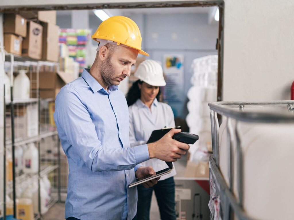 Mężczyzna w kasku ochronnym skanuje tag RFID za pomocą mobilnego czytnika w magazynie, obok niego kobieta z tabletem.