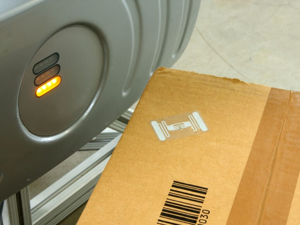 Zbliżenie na karton z naklejonym tagiem RFID i kodem kreskowym, umieszczony obok anteny RFID z włączoną sygnalizacją świetlną.